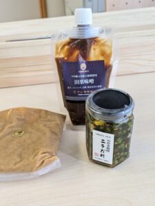 Assorted Japanese pantry items on a light wood table: a brown miso sauce pouch, a jar of pickled vegetables, and a plastic-wrapped item nearby.