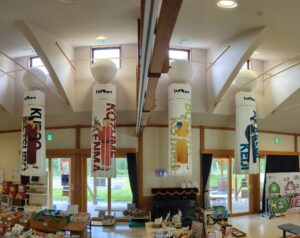 Inside a shop or exhibition space with tall vertical banners featuring anime characters and large white lanterns hanging from the ceiling.